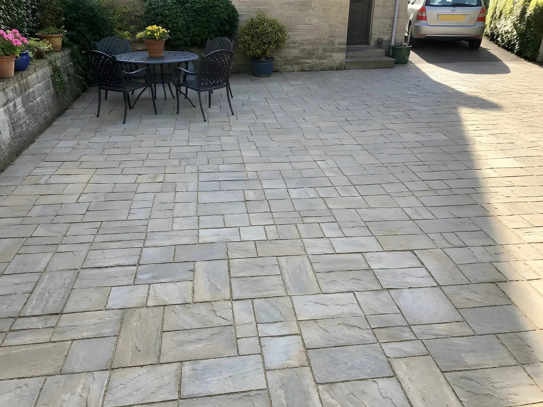 La même cour aux dalles grises et beiges propres, joints nets, même salon de jardin en fer devant la maison en pierre