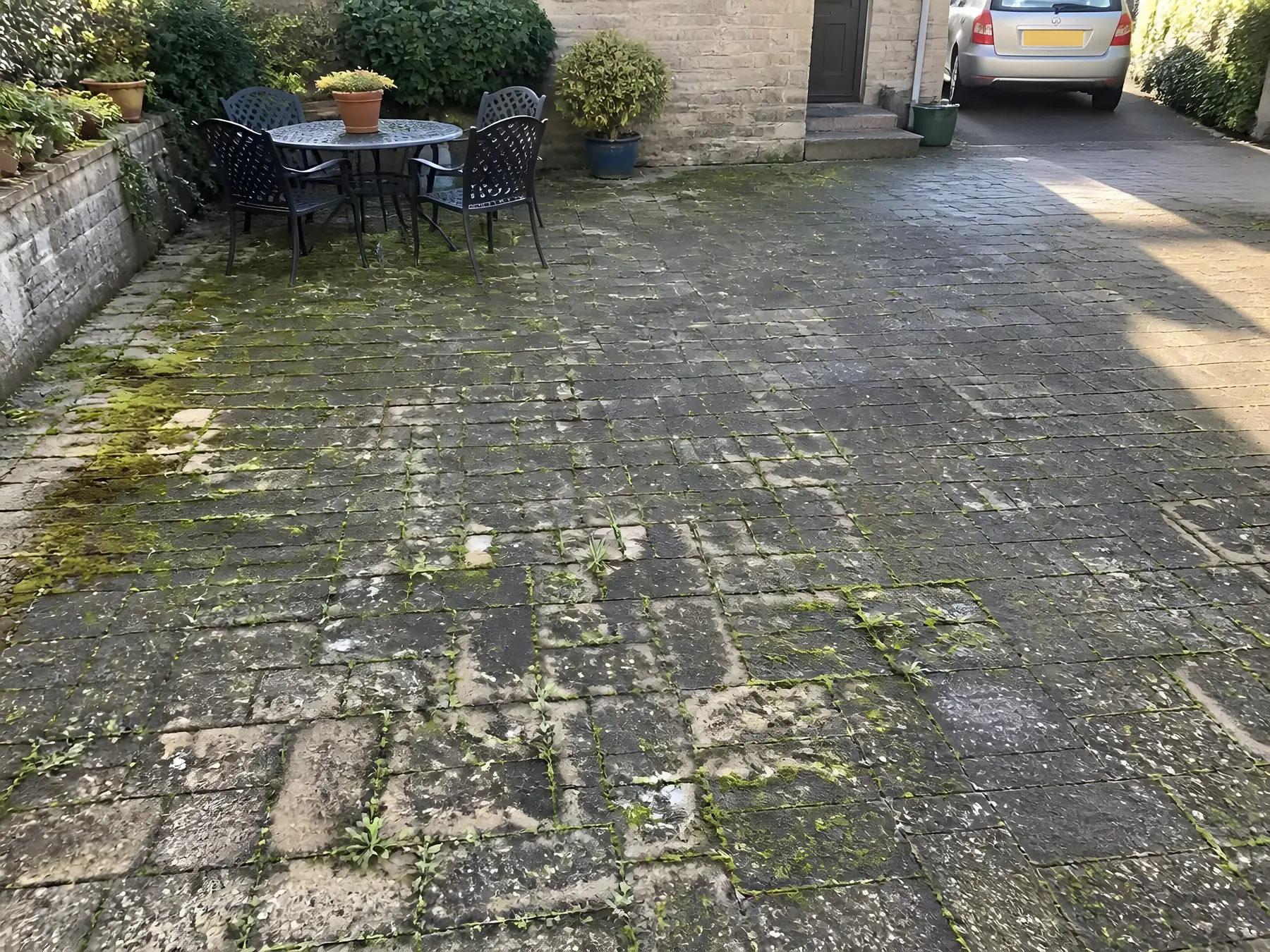 Cour dallée noircie, mousse verte et herbe dans les joints, salon de jardin en fer devant une maison en pierre