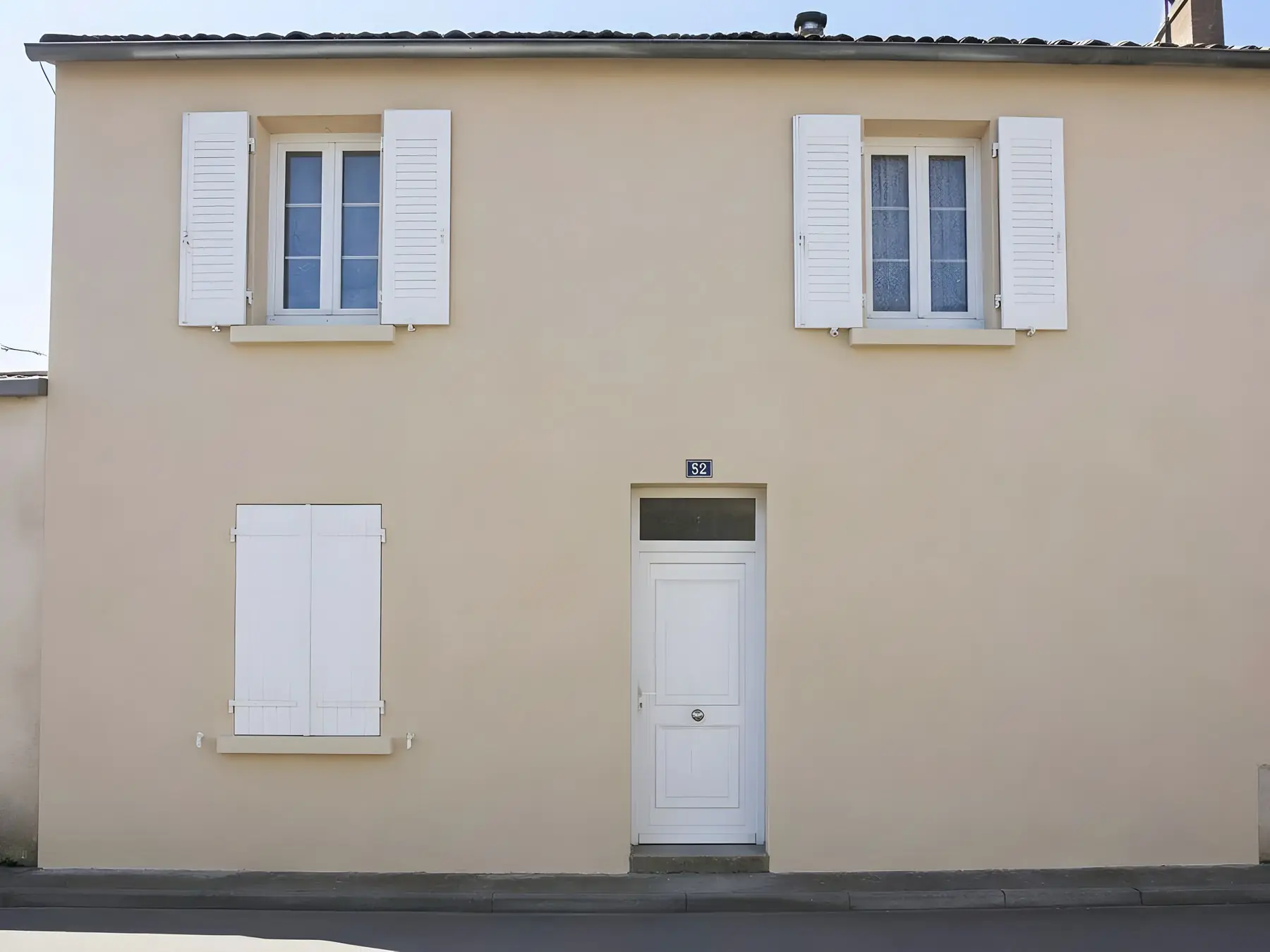 La même façade sur rue, enduit beige propre et uniforme, sans coulure, mêmes volets et même porte blancs
