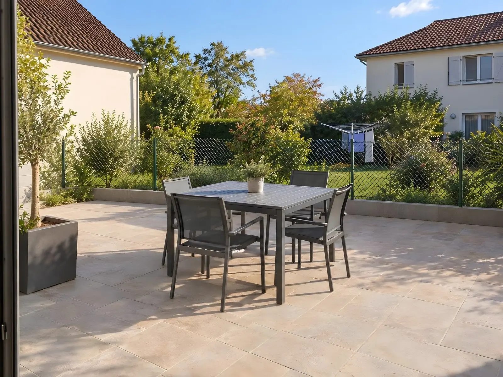 Terrasse dallée, table et quatre chaises, grillage bas ouvrant sur le jardin voisin, son étendoir et sa maison